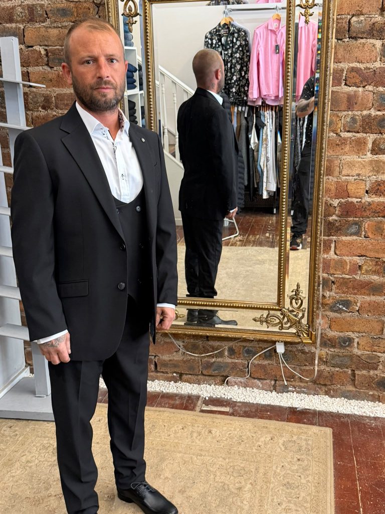 Customer wearing a black three-piece suit and white shirt beside the fitting-room mirror.