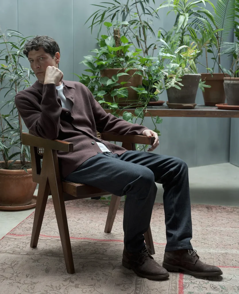 Model in brown knitwear and dark trousers seated by a window