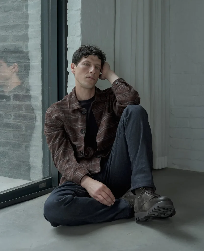 Model wearing a brown jacket seated beside a window